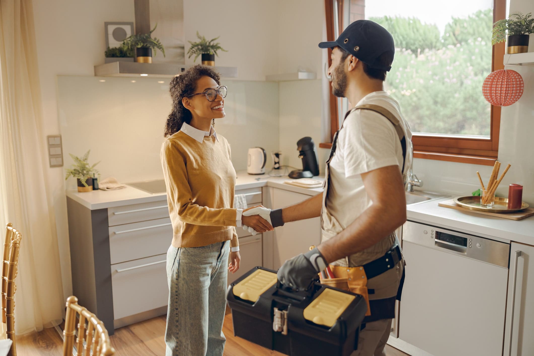 A woman in a kitchen shakes hands with a handyman from Armstrong Plumbing, who carries a toolbox and is ready to handle plumbing repairs.