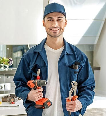 A man wearing a blue work uniform and cap stands indoors, smiling. He grips a cordless drill in one hand and pliers in the other, ready to tackle plumbing repairs with expertise.